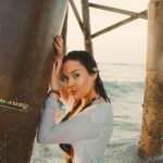 Isadora in Badebekleidung und offener Bluse an einem Holzsteg am Meer, warmes Abendlicht