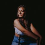 Vivienne in hellem Top und Jeans sitzend, warmes Streiflicht vor dunklem Hintergrund
