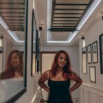 Noemi in schwarzem Kleid in einem Flur mit Spiegel, warmes Kunstlicht, rotes Haar