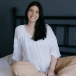Sienna in weißer Bluse entspannt auf einem Bett sitzend, helles ruhiges Zimmer